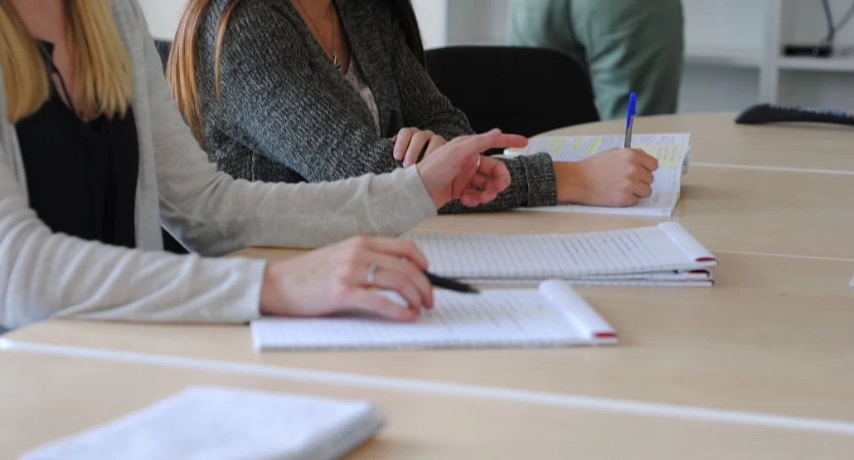 Uczniowie siedzący przy stole podczas zajęć, zapisujący notatki w zeszytach i wskazujący fragmenty materiału w podręczniku. Ujęcie pokazuje pracę nad zadaniami i wspólne omawianie treści, charakterystyczne dla systematycznego przygotowania do matury w Kielcach z Symposio.